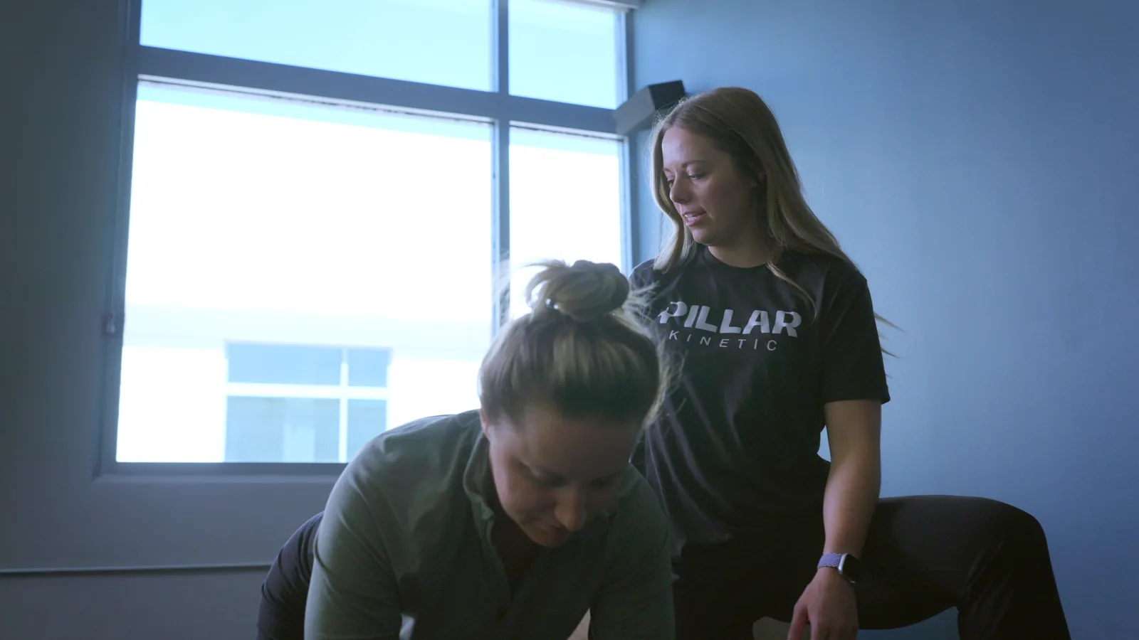 A physical therapist watching a patient's hips as she lowers into a hinge in the clinic gym