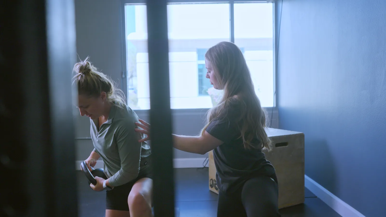 A physical therapist steadying a woman as she lifts a kettlebell beside a box in the clinic gym