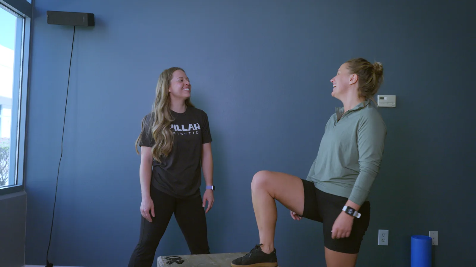 A patient with one foot up on a box, laughing with her physical therapist in the clinic's exercise room