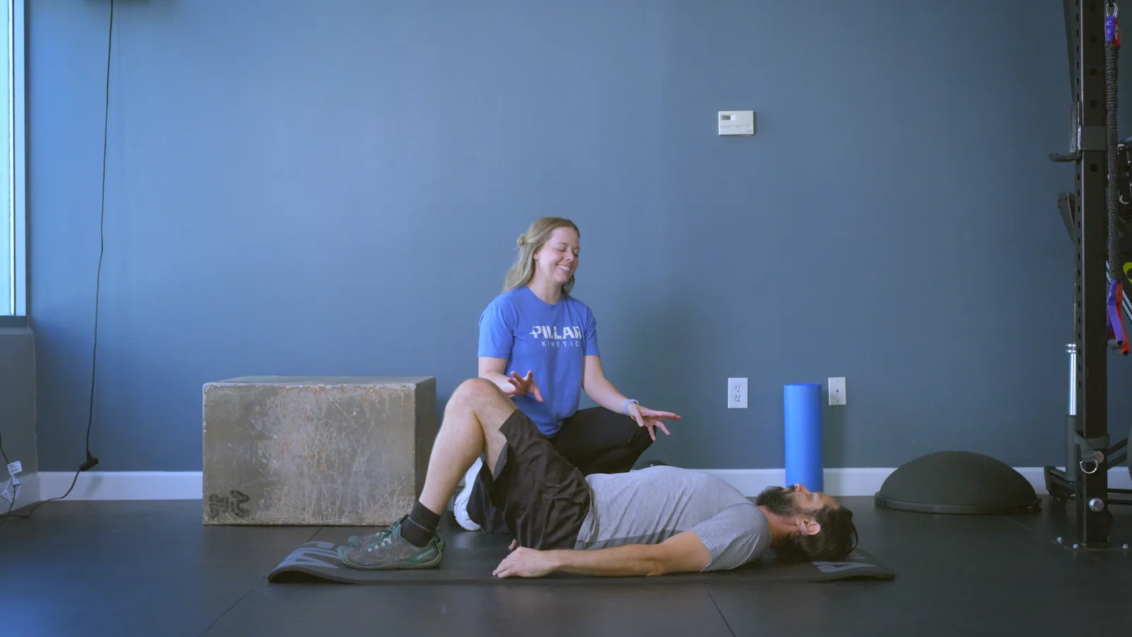 A patient lying on a mat with his knees bent while his physical therapist kneels behind him, explaining