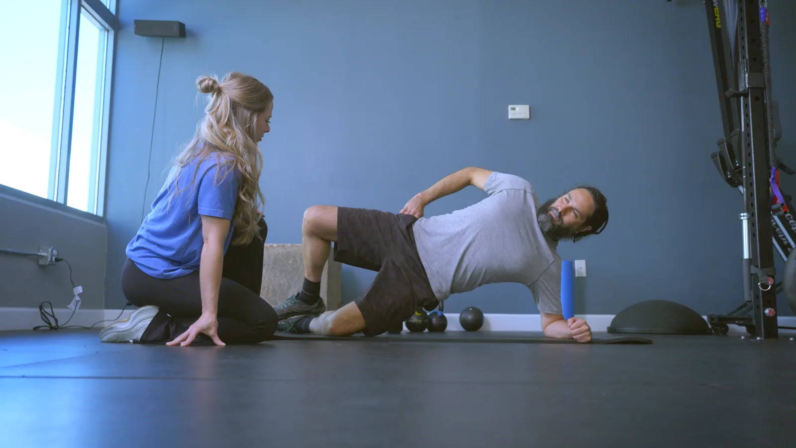 A physical therapist kneeling on a mat as she coaches a man through a side-lying hip exercise
