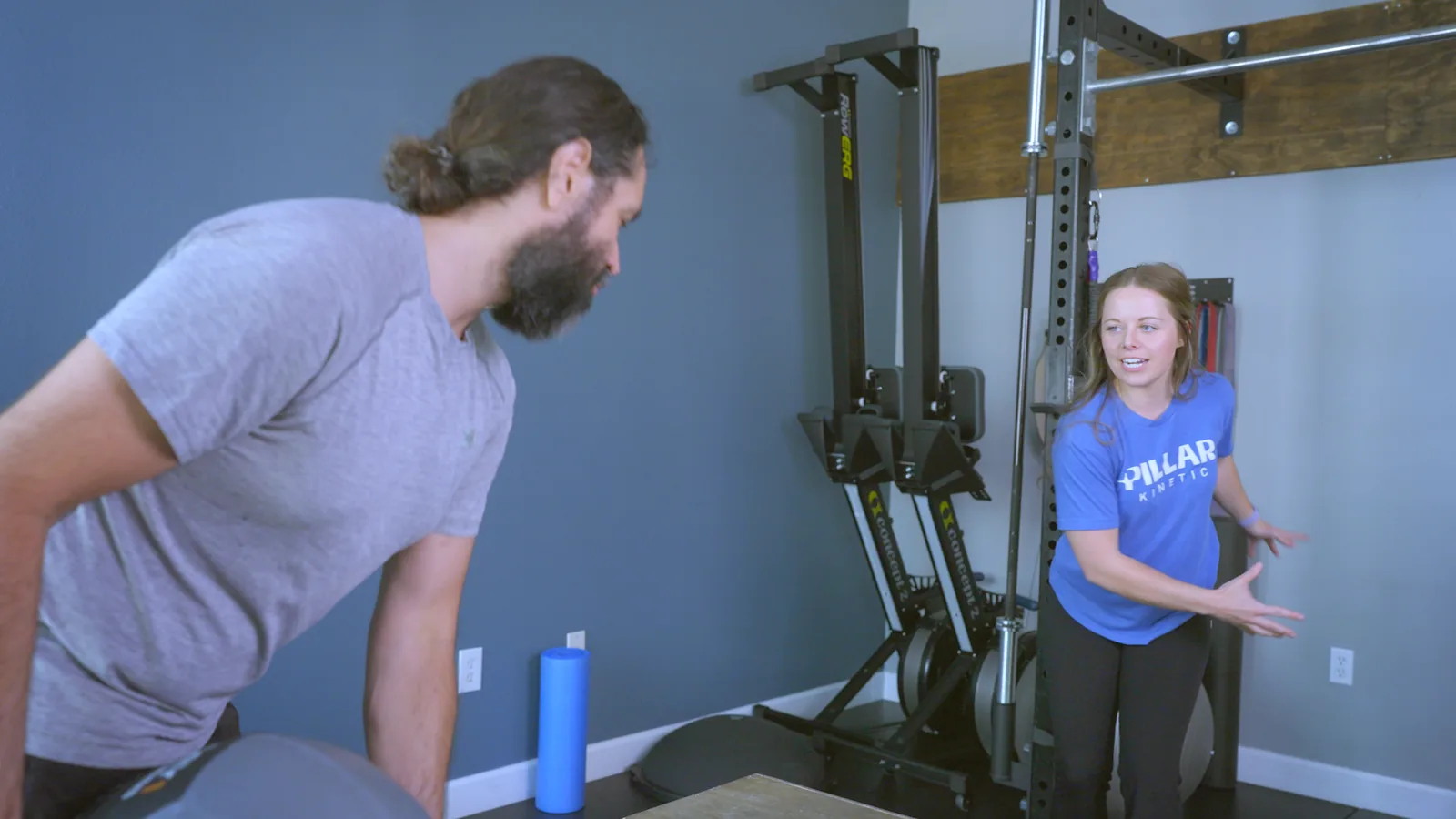 A man hinging forward in the clinic gym while a physical therapist demonstrates the movement beside the rack