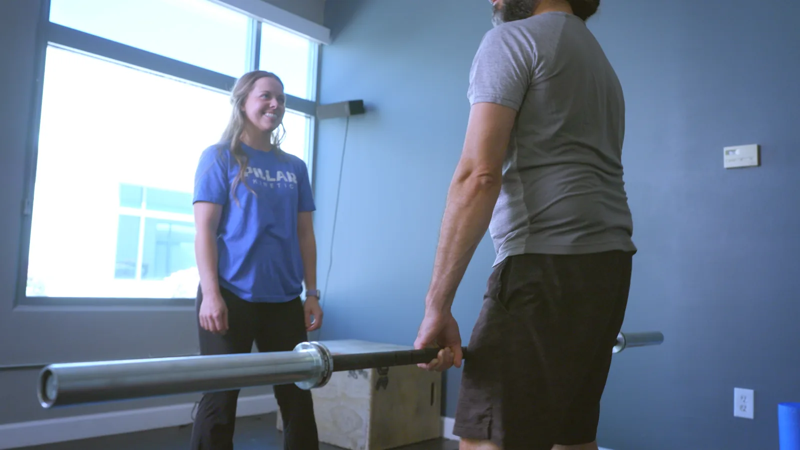A man gripping a barbell at his hips while his physical therapist coaches him in the clinic gym