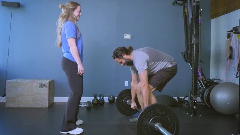 A physical therapist watching a man set his grip on a loaded barbell at the start of a deadlift