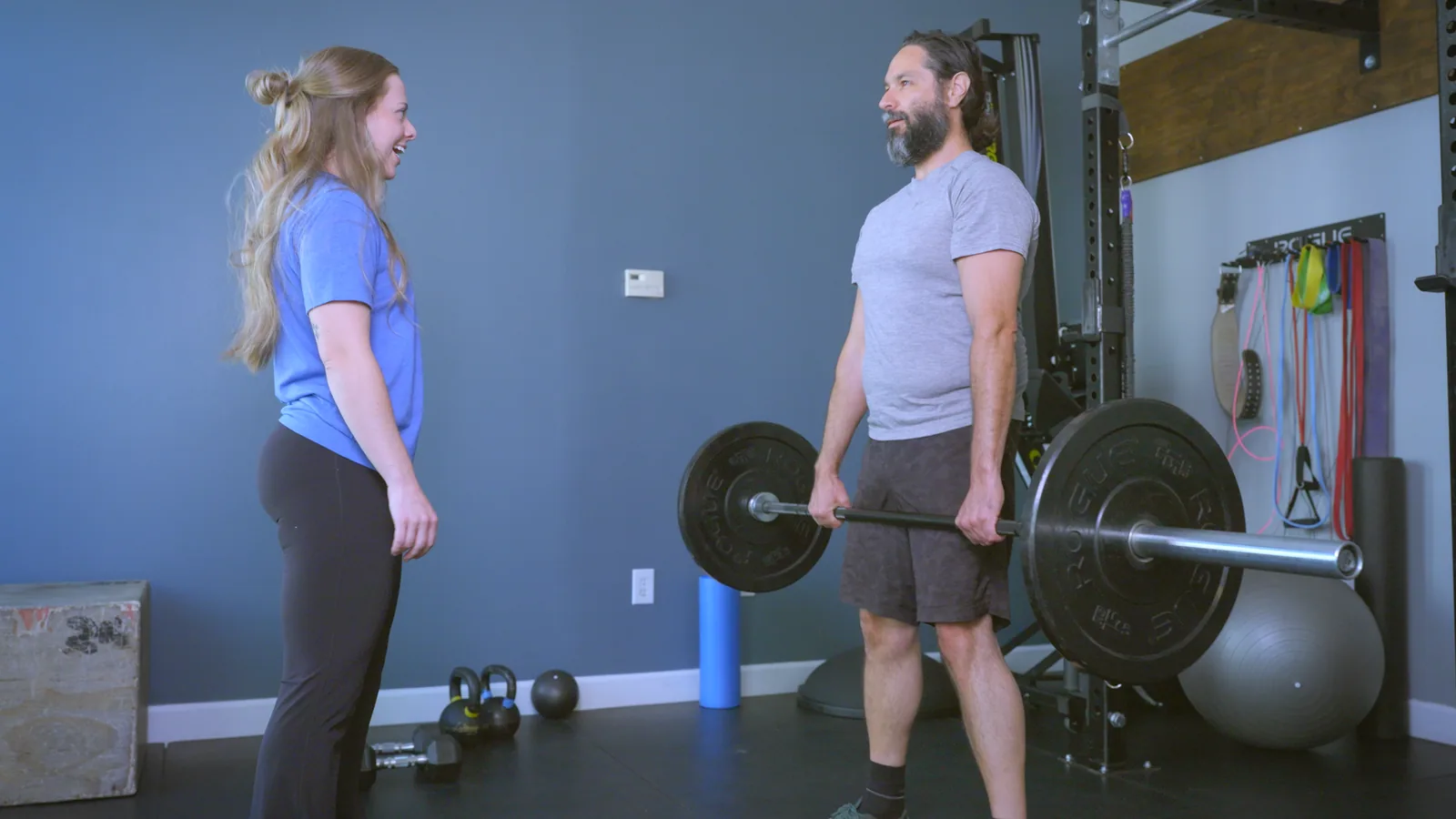 A man holding a loaded barbell at his hips as his physical therapist looks on