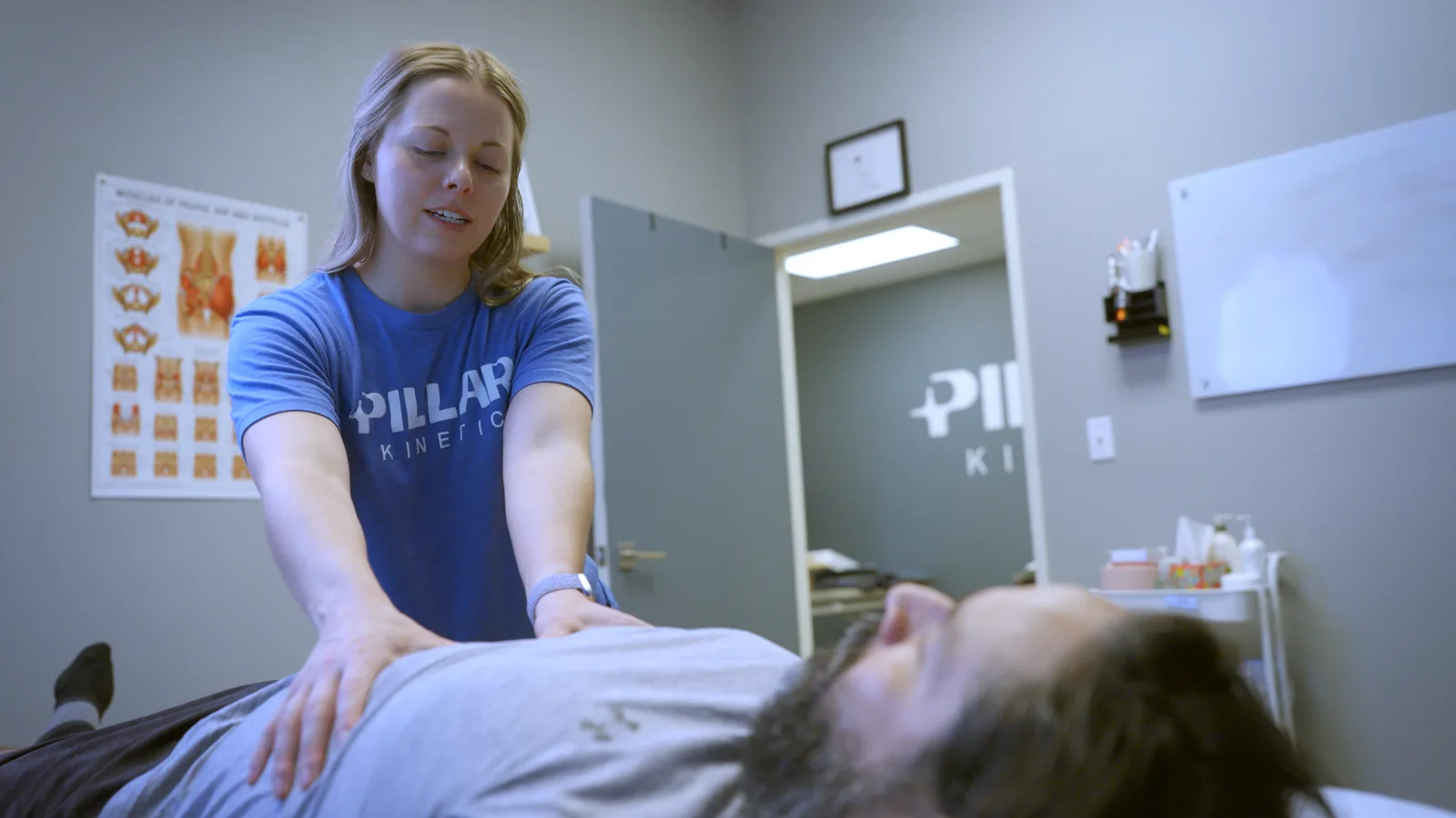 A physical therapist assessing a male patient on the treatment table, a chart of the pelvic muscles on the wall behind her
