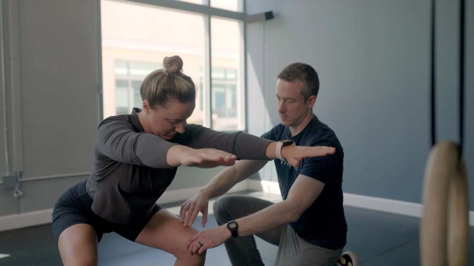 A physical therapist kneeling beside a woman in a split stance as she reaches both arms forward